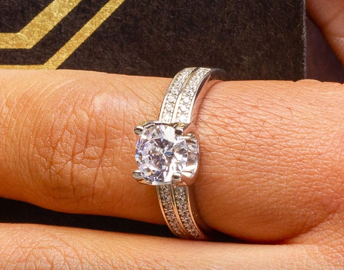 Sterling silver solitaire ring with a double row of pavé stones on a finger, resting on a black and gold box